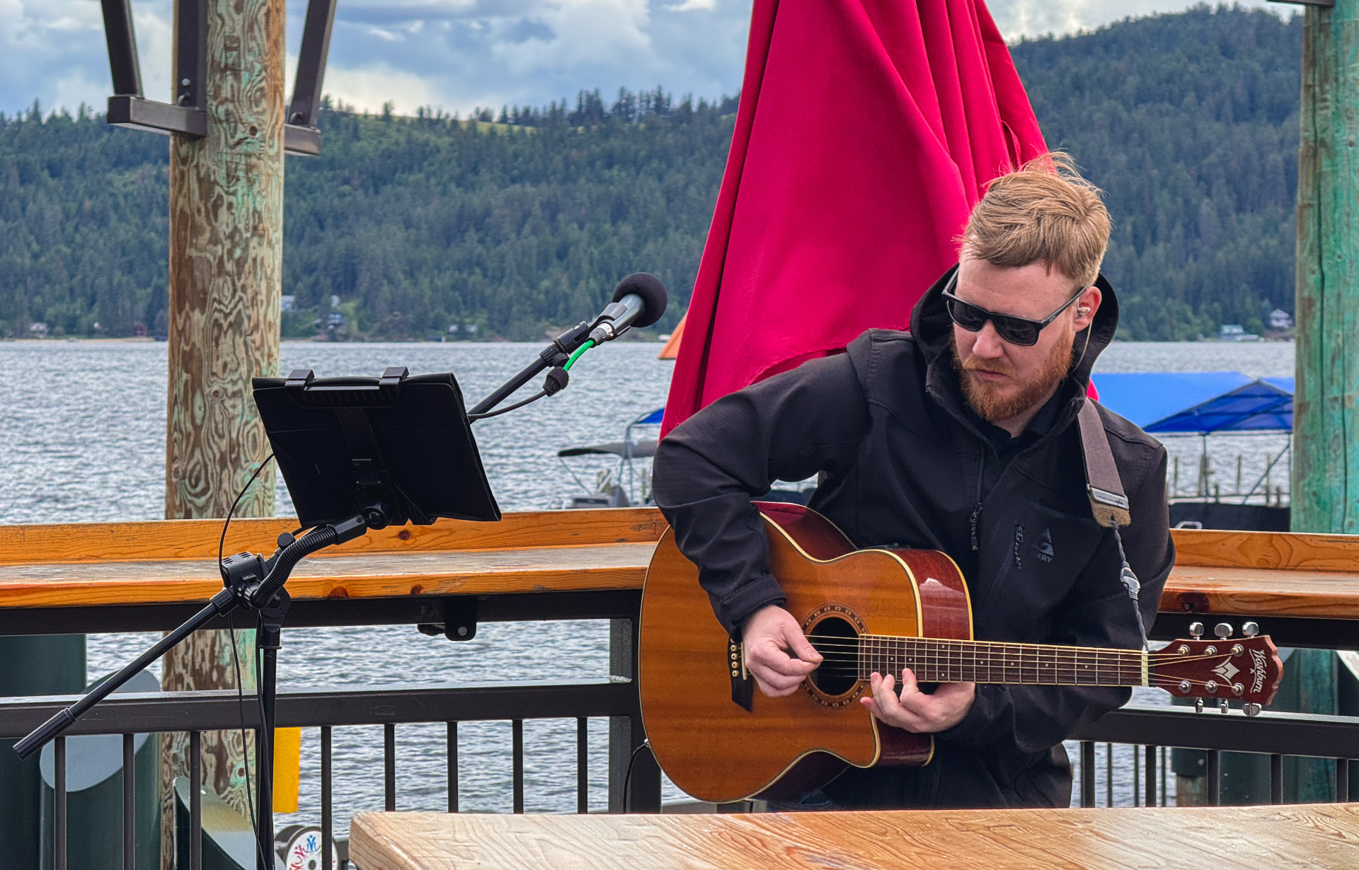 Acoustic musician performing on a lakeside patio in Coeur d'Alene with Lake Coeur d'Alene and forested hills in the background